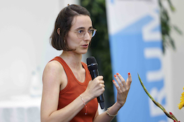 Die Referentin Janine Bürkli spricht in ein Handmikrofon und gestikuliert mit der linken Hand. Im Hintergrund ist das blauweisse Banner mit dem Forumslogo erkennbar.