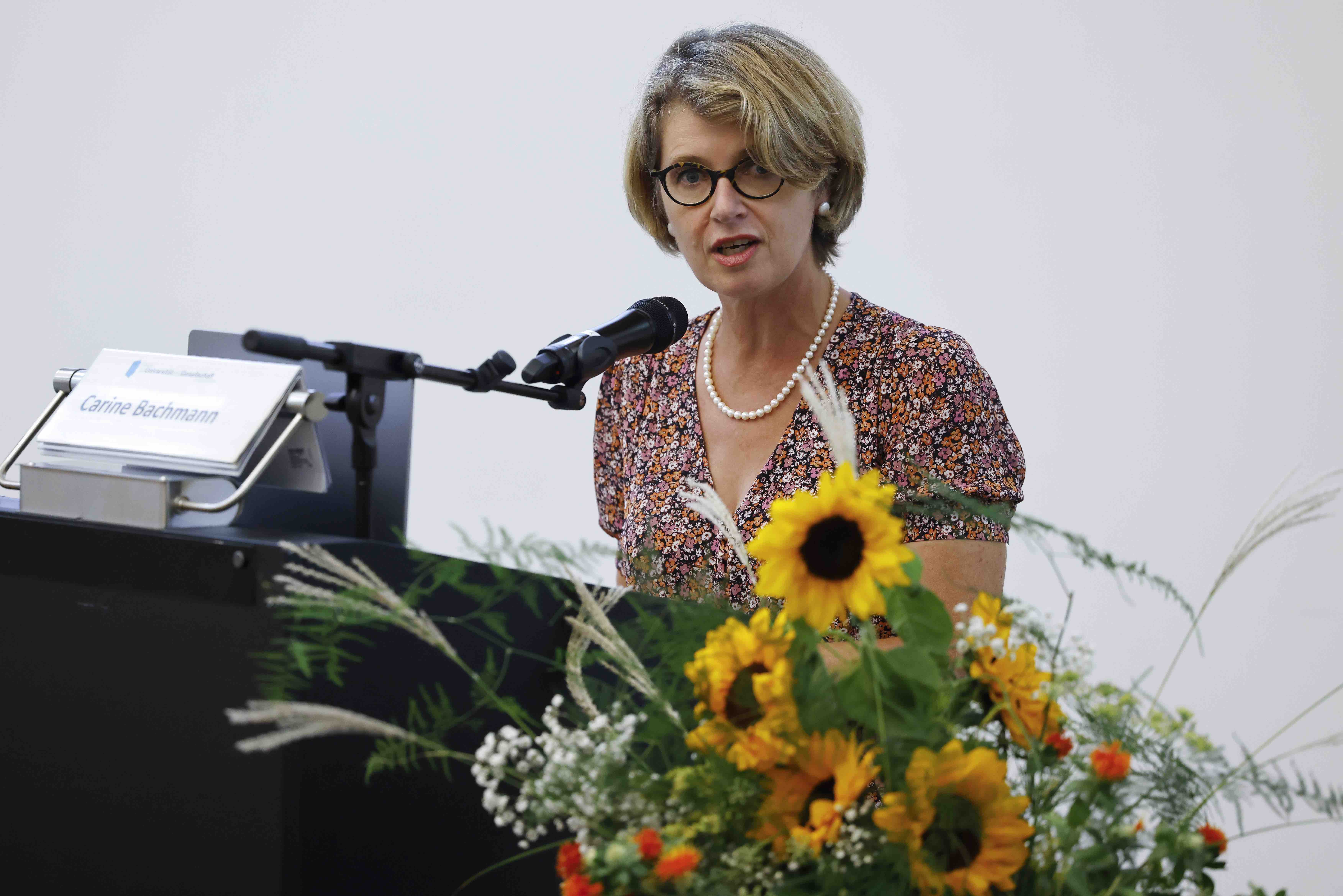 Die Referentin Carine Bachmann steht hinter einem Rednerpult. Sie trägt ein rotweiss geblümtes Kleid und eine Perlenkette. Neben ihr ist ein Blumenstrauss mit Sonnenblumen erkennbar.