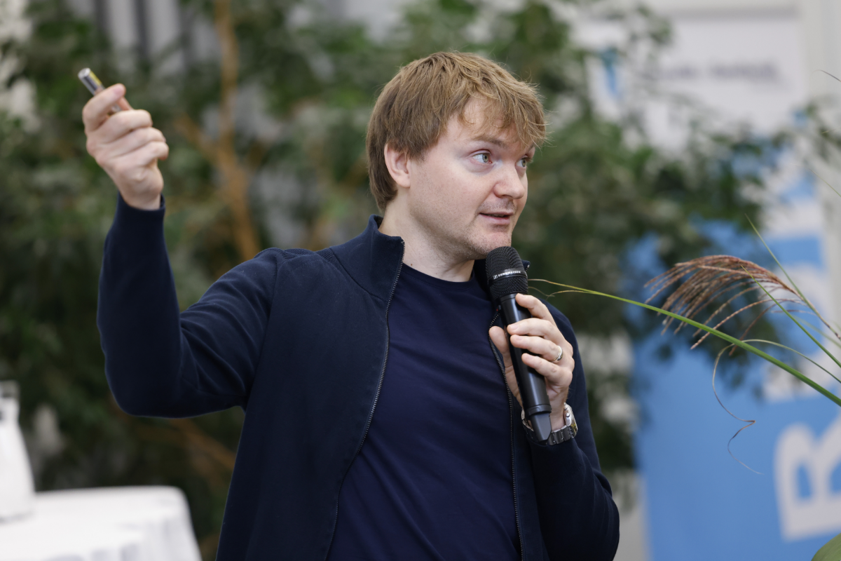 Ein Mann in dunkler Kleidung hält einen Vortrag über künstliche neuronale Netzwerke. In seiner linken Hand hält er ein Handmikrofon, mit der rechten, erhobenen Hand gestikuliert er. Im Hintergrund ist eine dunkelgrüne Zimmerpflanze erkennbar und ein Teil des Banners mit dem blau-weissen Forumslogo.