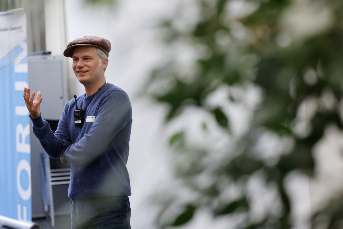 Ein Mann mit Schiebermütze und blauem Pullover lacht und gestikuliert mit der rechten Hand. Er hält einen Vortrag über maschinelles Lernen. Rechts im Bild ist eine grüne Zimmerpflanze erkennbar, am linken Rand ein Teil des blau-weissen Logos des Forums für Universität und Gesellschaft.