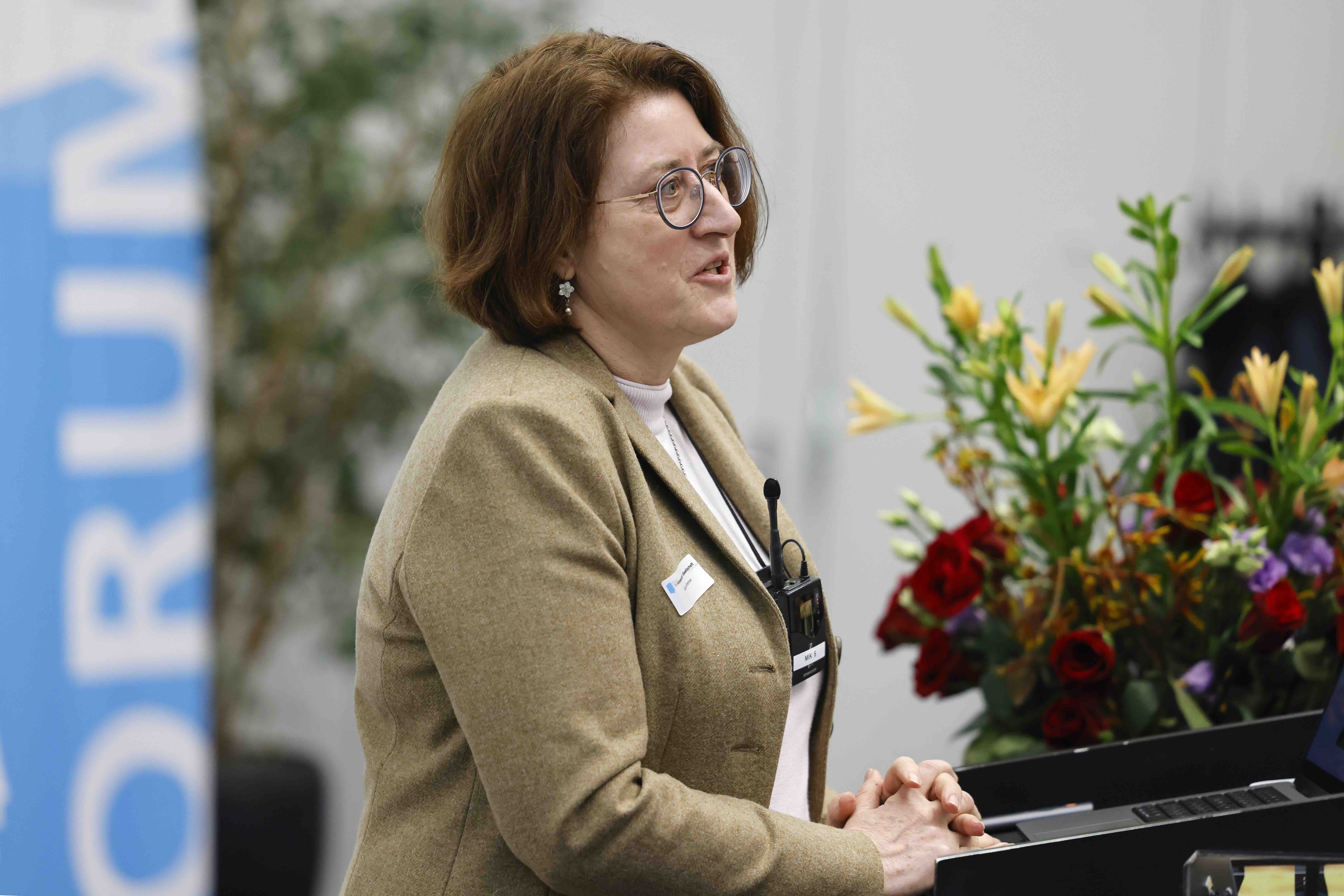 Eine Frau ist am Sprechen. Sie trägt ein Beiges Oberteil und ein Umhängemikrofon um den Hals. Neben ihr steht ein bunter Blumenstrauss und im Hintergrund ist das blau-weisse Banner des Forums für Universität und Gesellschaft erkennbar.