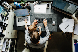 Die Arbeitswelt im Wandel: Sicht von oben auf einen Menschen, der mit  Kind im Homeoffice an einem Schreibtisch sitzt und am Computer arbeitet.