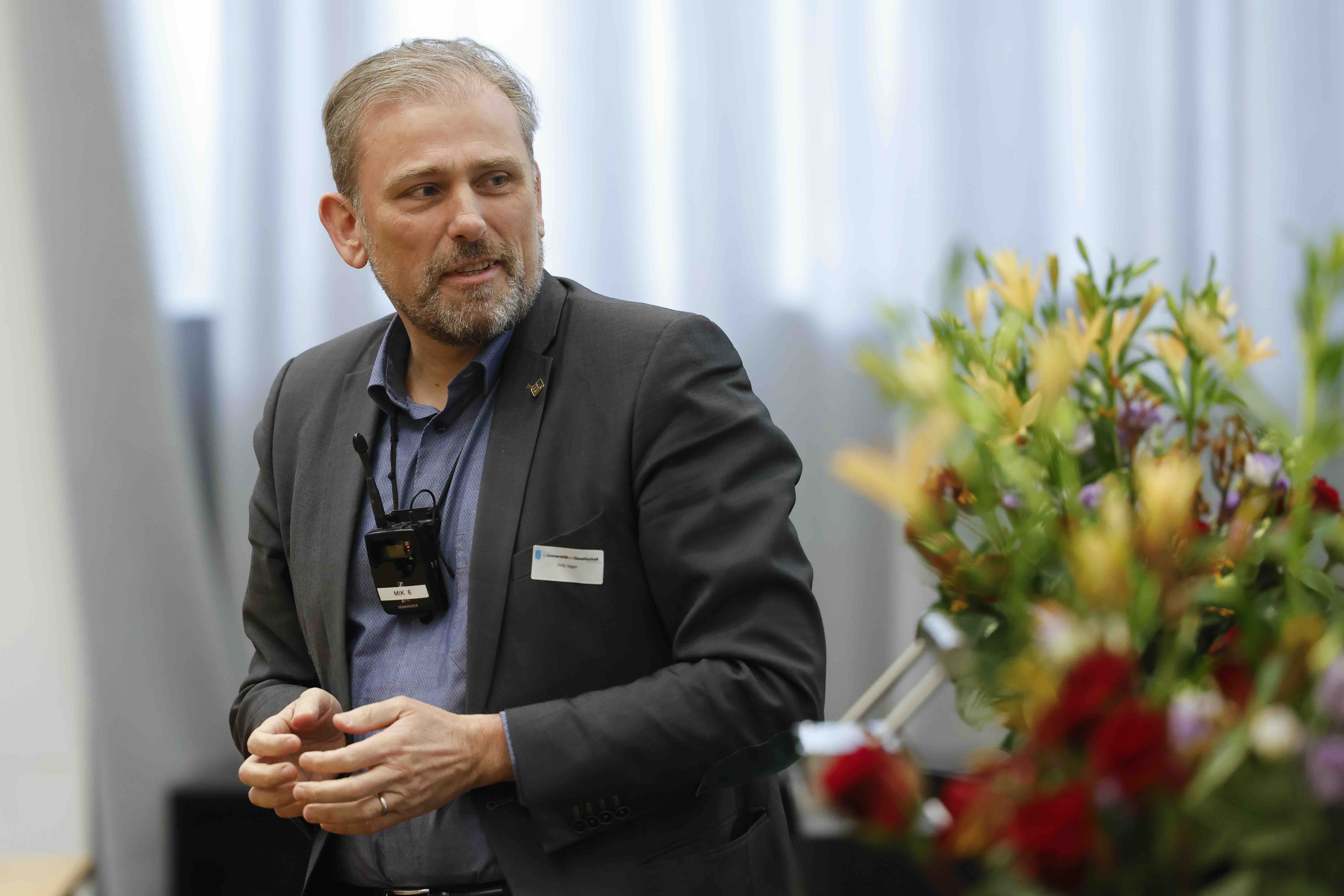 Ein Mann in einem grauen Jackett und blauen Hemd trägt um den Hals ein Umhängemikrofon. Seine Hände berühren sich und neben ihm steht ein Blumenstrauss