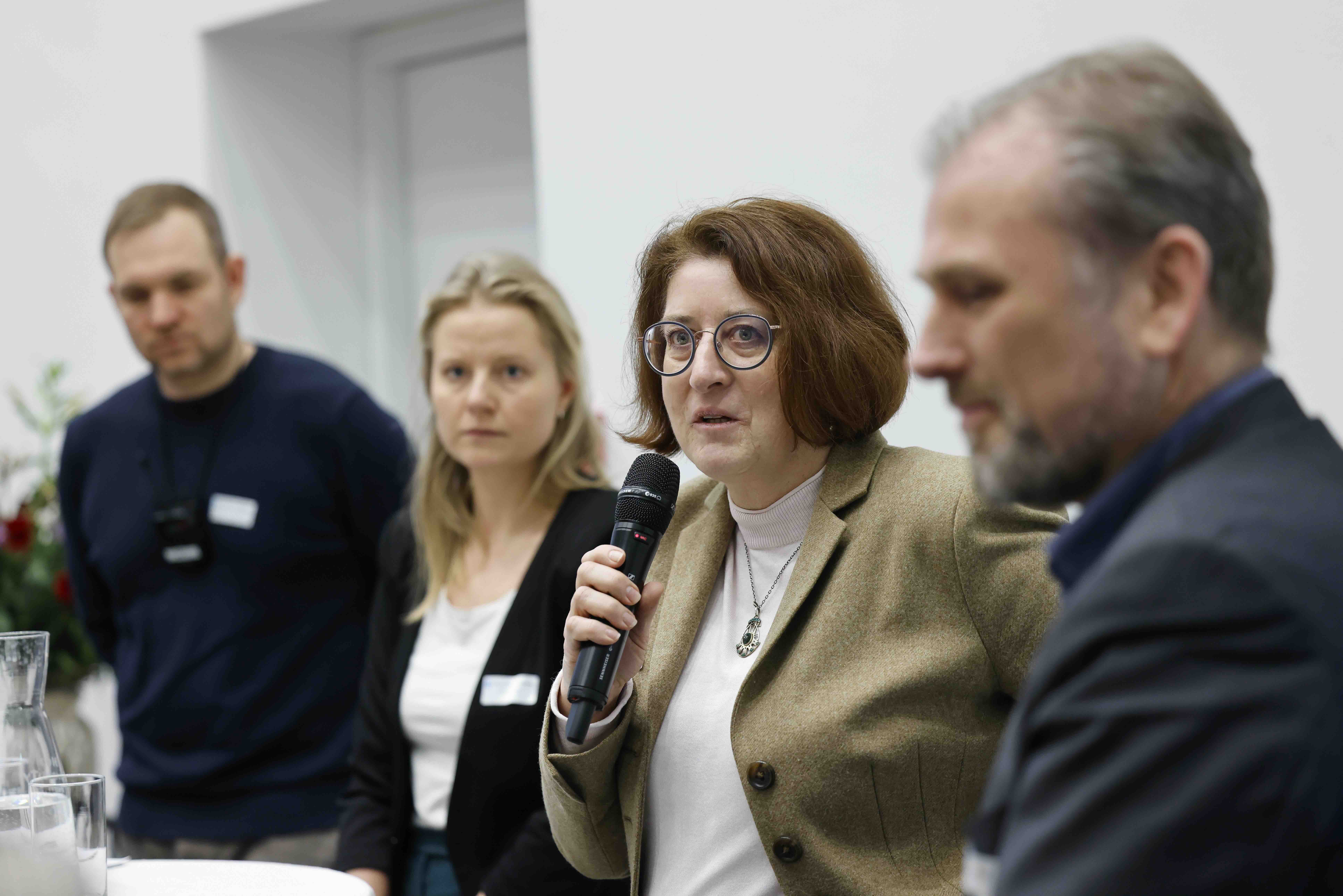 Vier Personen stehen nebeneinander. Die zweite Person von rechts spricht in ihr Handmikrofon.