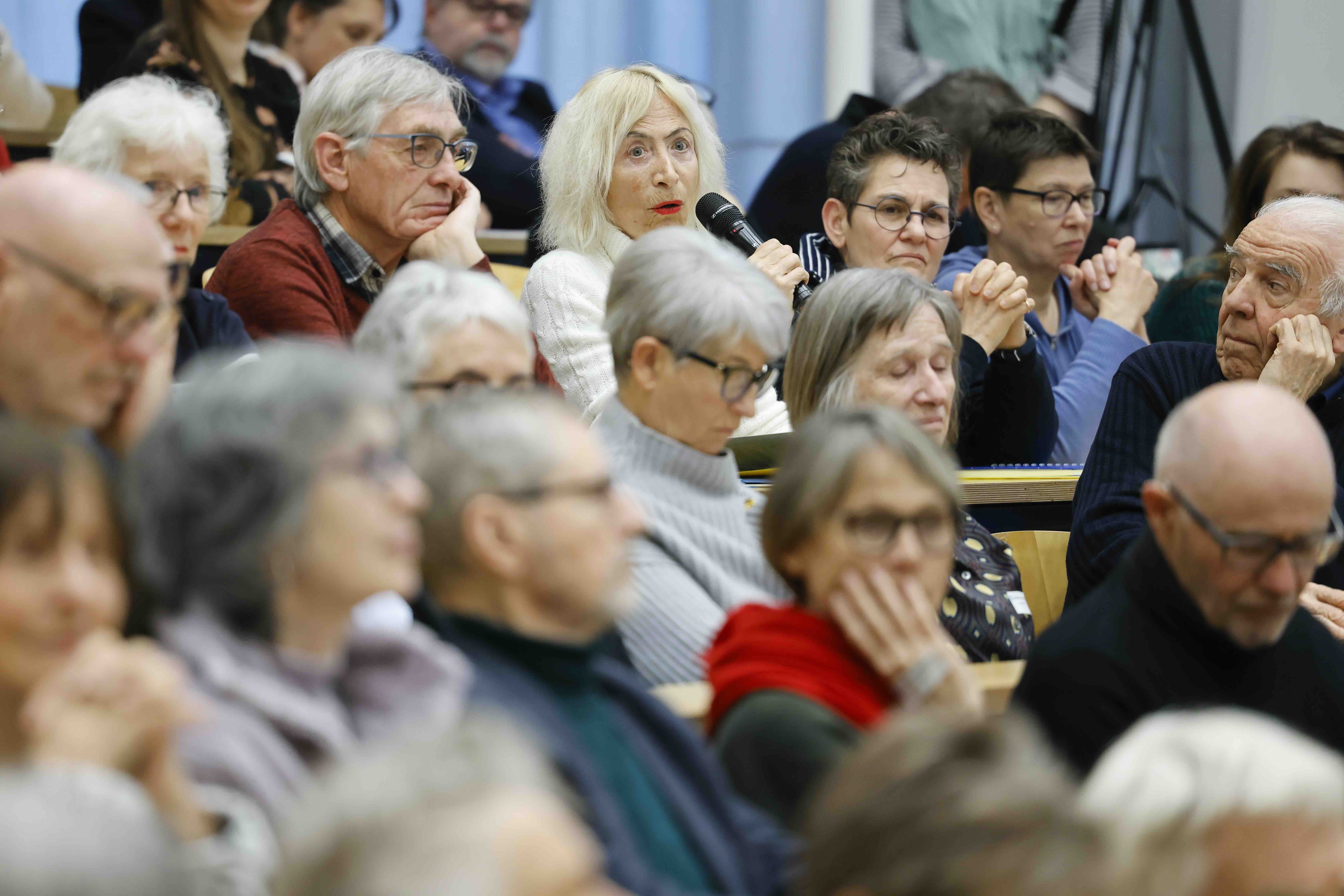 Menschen sitzen in einem Hörsaal. Sie hören einer Frau mit blonden Haaren zu, die in ein Handmikrofon spricht.