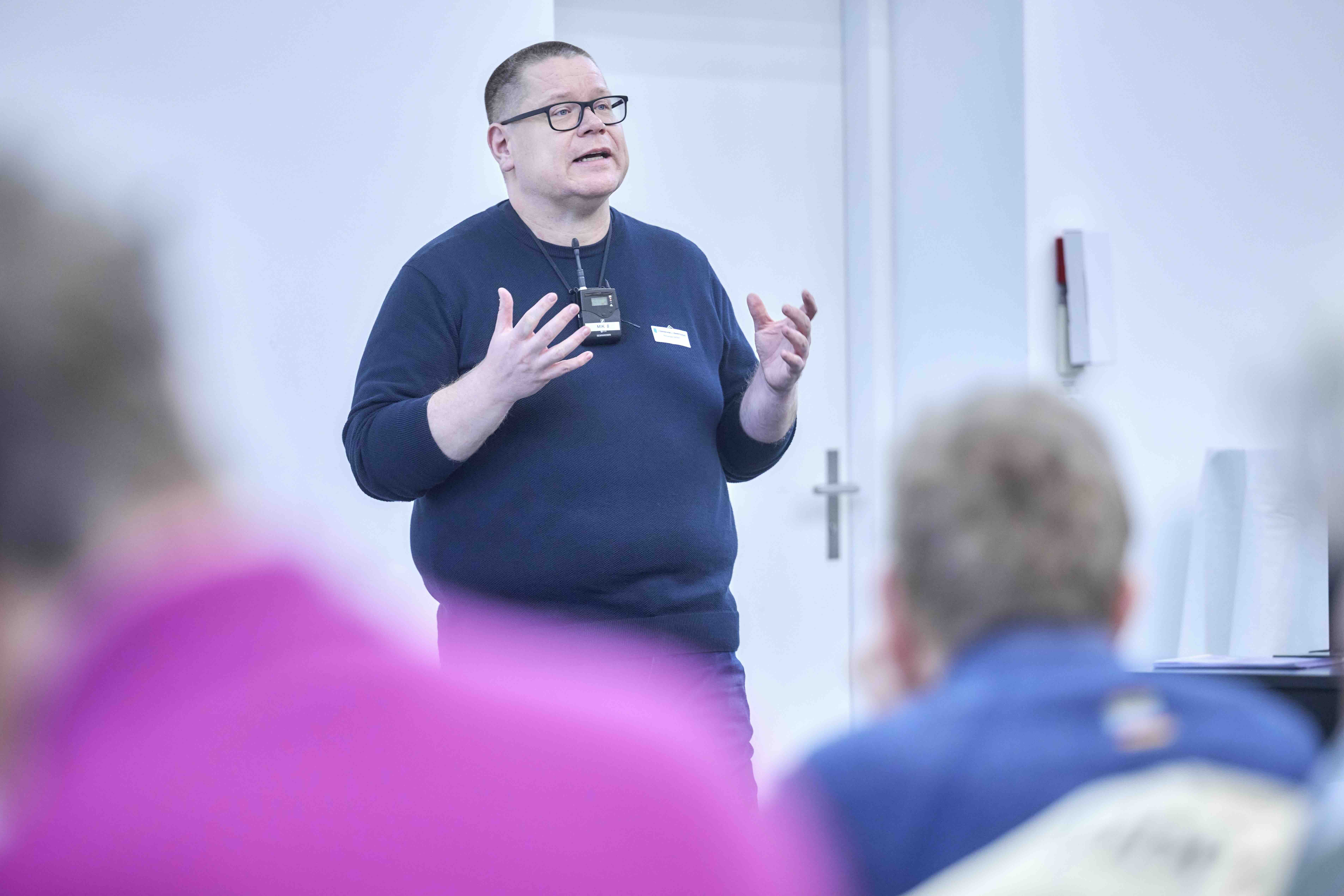 Ein Mann steht vor Publikum und gestikuliert mit beiden Händen. Er trägt dunkle Kleidung, eine Brille und um den Hals ein Umhängemikrofon. Im Vordergrund sind Personen aus dem Publikum von hinten erkennbar.