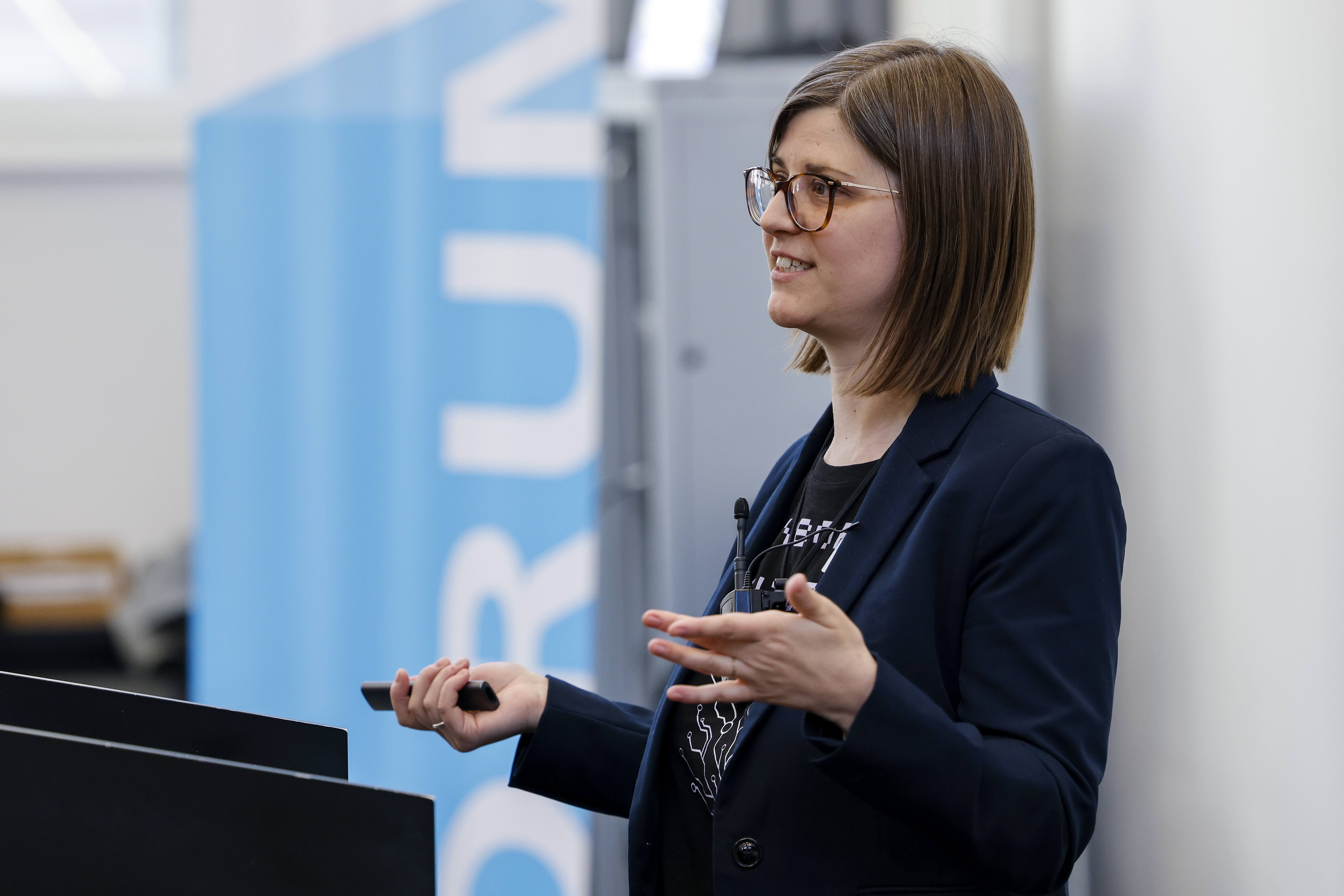 Jennifer Scurrell hält ein Referaten gestikuliert mit beiden Händen. Sie trägt dunkle Kleidung und eine Brille. in der rechten Hand hält sie einen Pointer. Im Hintergrund ist ein Banner mit dem blau-weissen Forumslogo erkennbar.