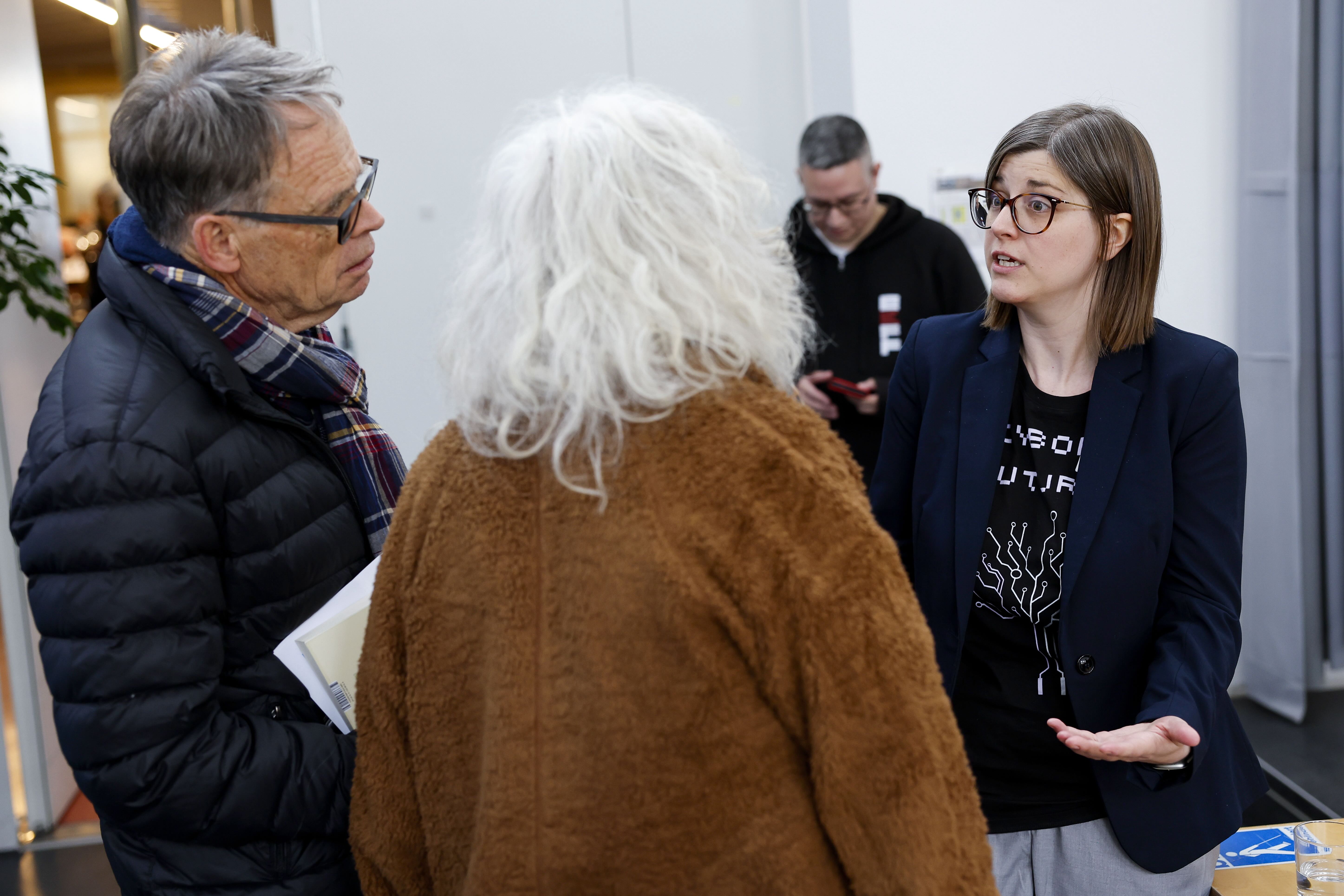 Ein Mann und eine Frau (er trägt eine schwarze, sie eine braune Jacke) diskutieren mit einer jungen Frau mit Brille und dunkler Kleidung. Im Hintergrund ist ein weiterer Mann erkennbar.