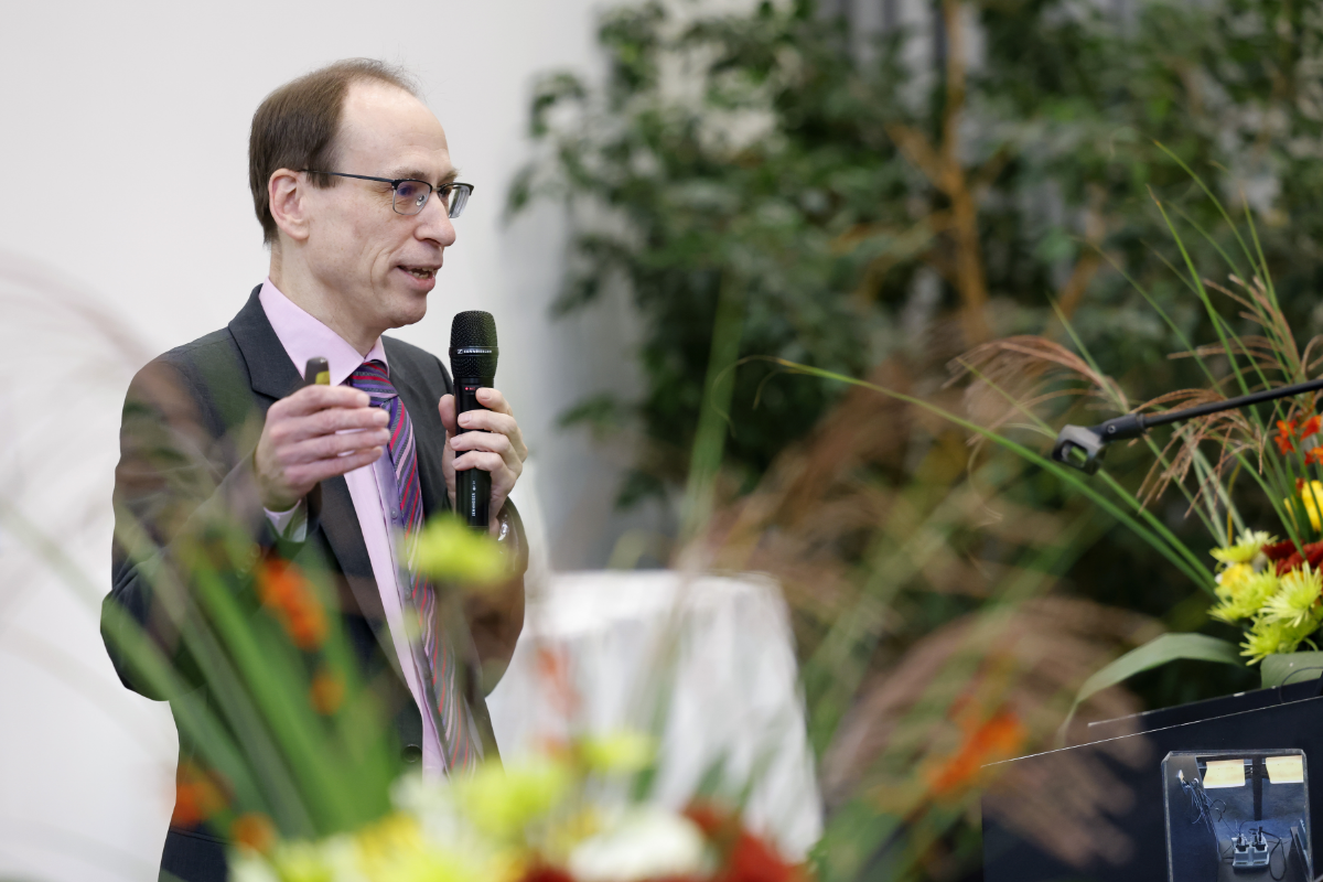 Claus Beisbart spricht in ein Handmikrofon und gestikuliert mit der einen Hand. Im Vordergrund ist ein Blumenstrauss teilweise erkennbar, im Hintergrund steht eine grüne Zimmerpflanze. Beisbart trägt einen grauen Anzug und ein rosa Hemd mit Kravatte.