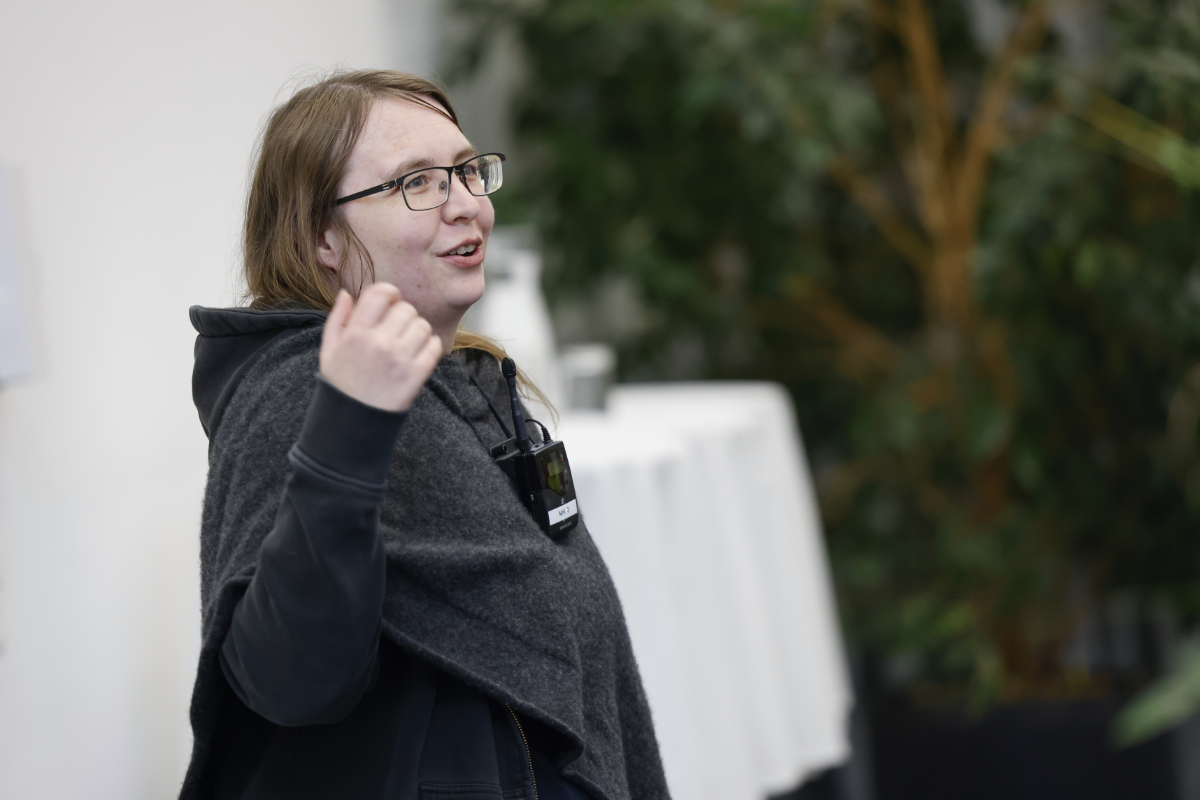 Petra Müller hält einen Vortrag über Bildbearbeitung und künstliche Intelligenz. Sie trägt eine Brille, schwarze Kleidung und einen grauen Umhang. In ihrer rechten Hand hält sie den Pointer, im Hintergrund ist eine grüne Zimmerpflanze erkennbar.