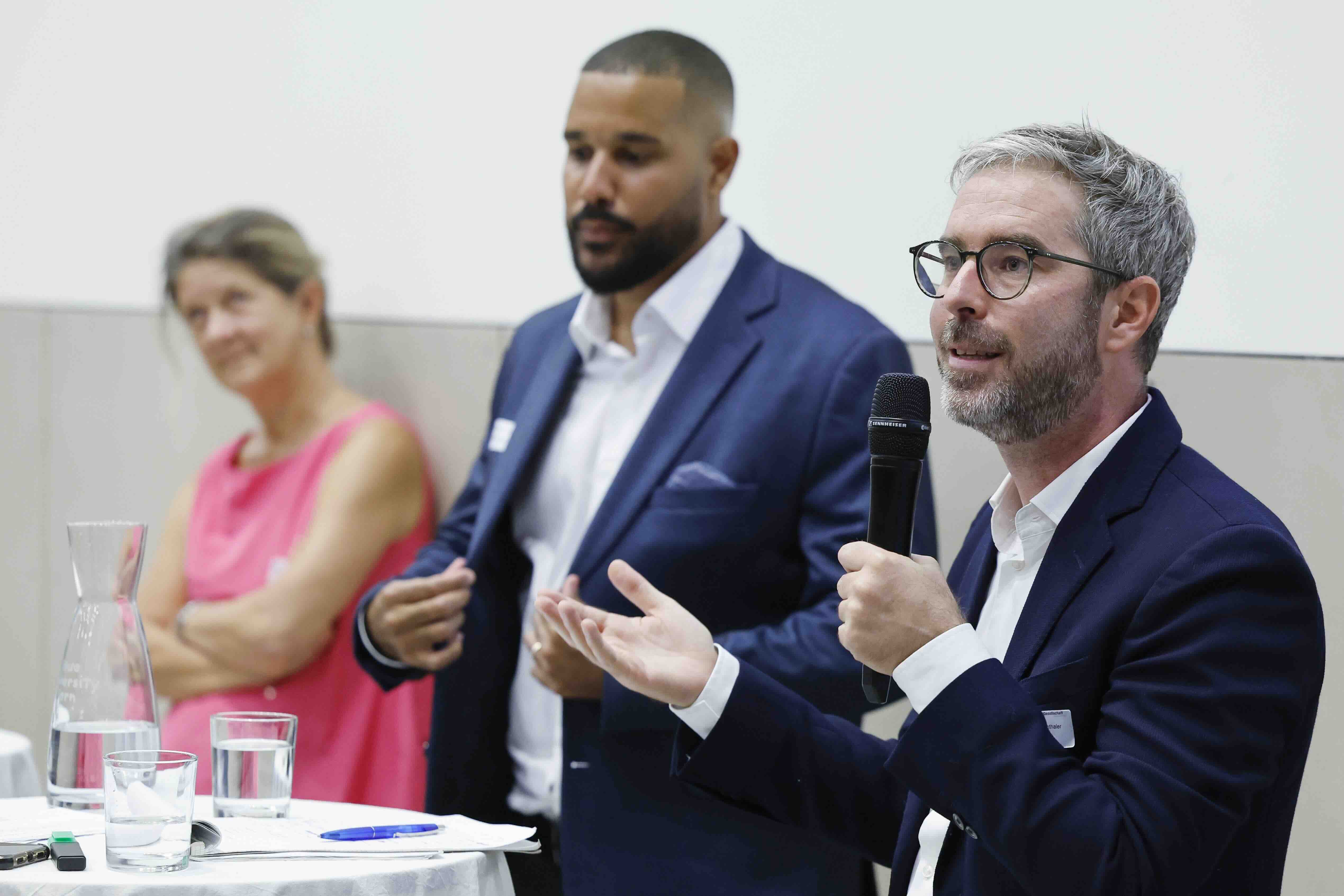 Michael Siegenthaler, Malik Hashim und Gudela Grote stehen hinter einem Stehtisch. Michael Siegenthaler hält ein Handmikrofon in der linken Hand und gestikuliert mit der rechten Hand.