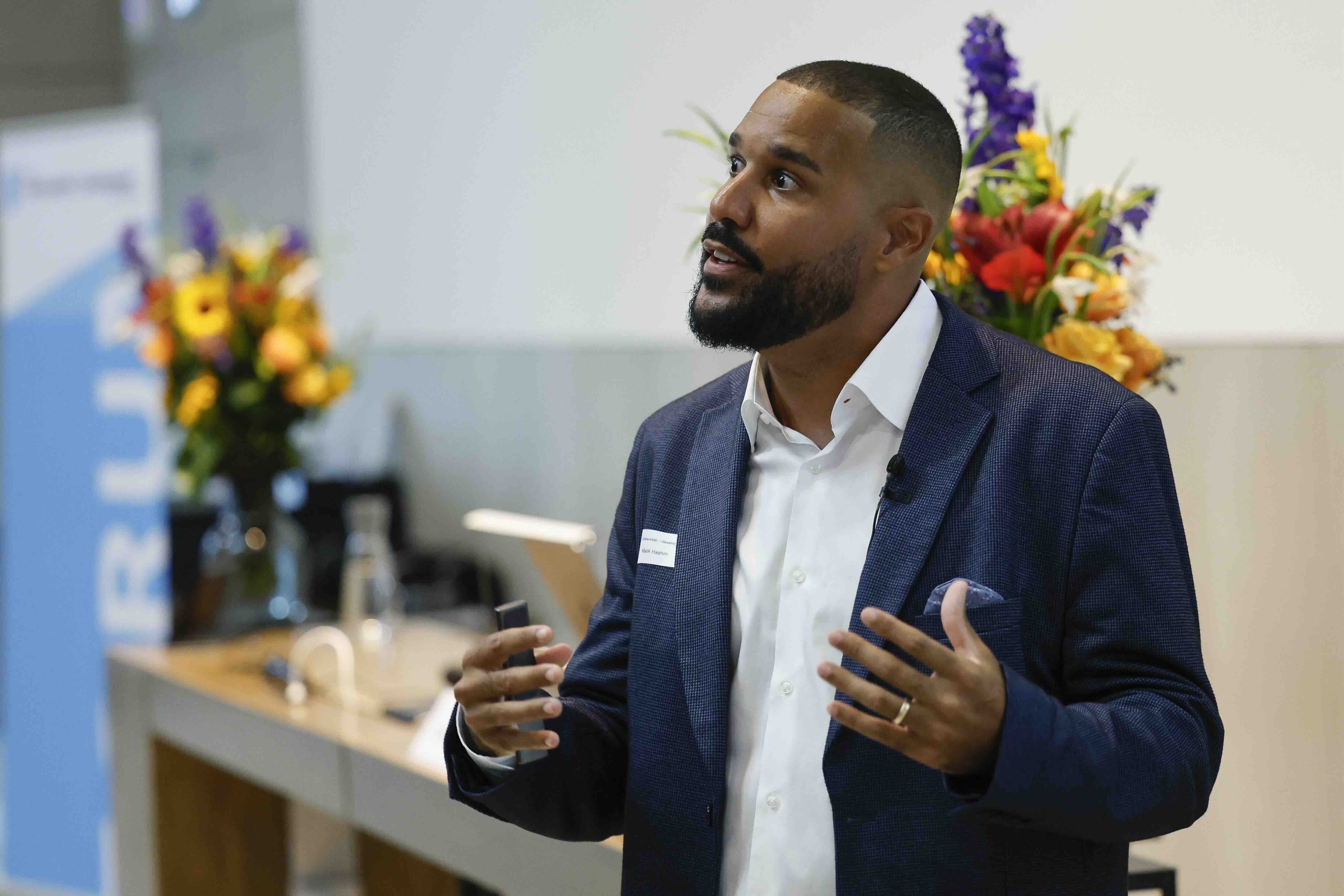 Malik Hashim von Kitoko People gestikuliert während seinem Referat mit beiden Händen. Hinter ihm steht ein Tisch mit zwei Blumensträussen, links im Bild ist ein Banner mit dem blau-weissen Logo des Forums für Universität und Gesellschaft zu sehen.