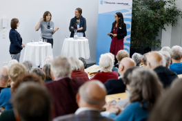 Vier Personen stehen während einer Podiumsdiskussion an zwei Stehtischen mit weissen Tischtüchern. Im Vordergrund ist ein Teil des Publikums sichtbar.